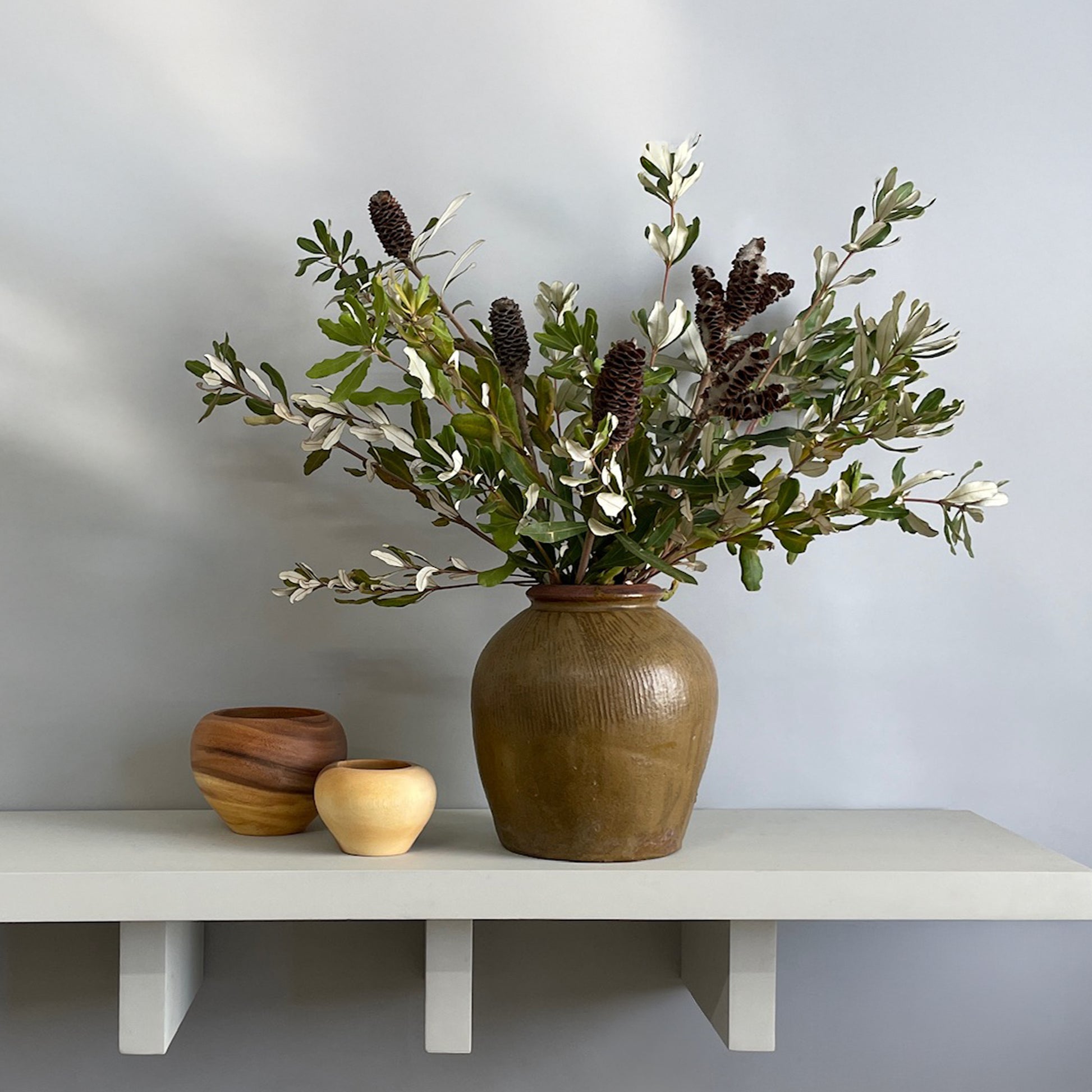 A large olive-green ginger jar with fresh integrifolia foliage styled on a decorative shelf with wooden accents.