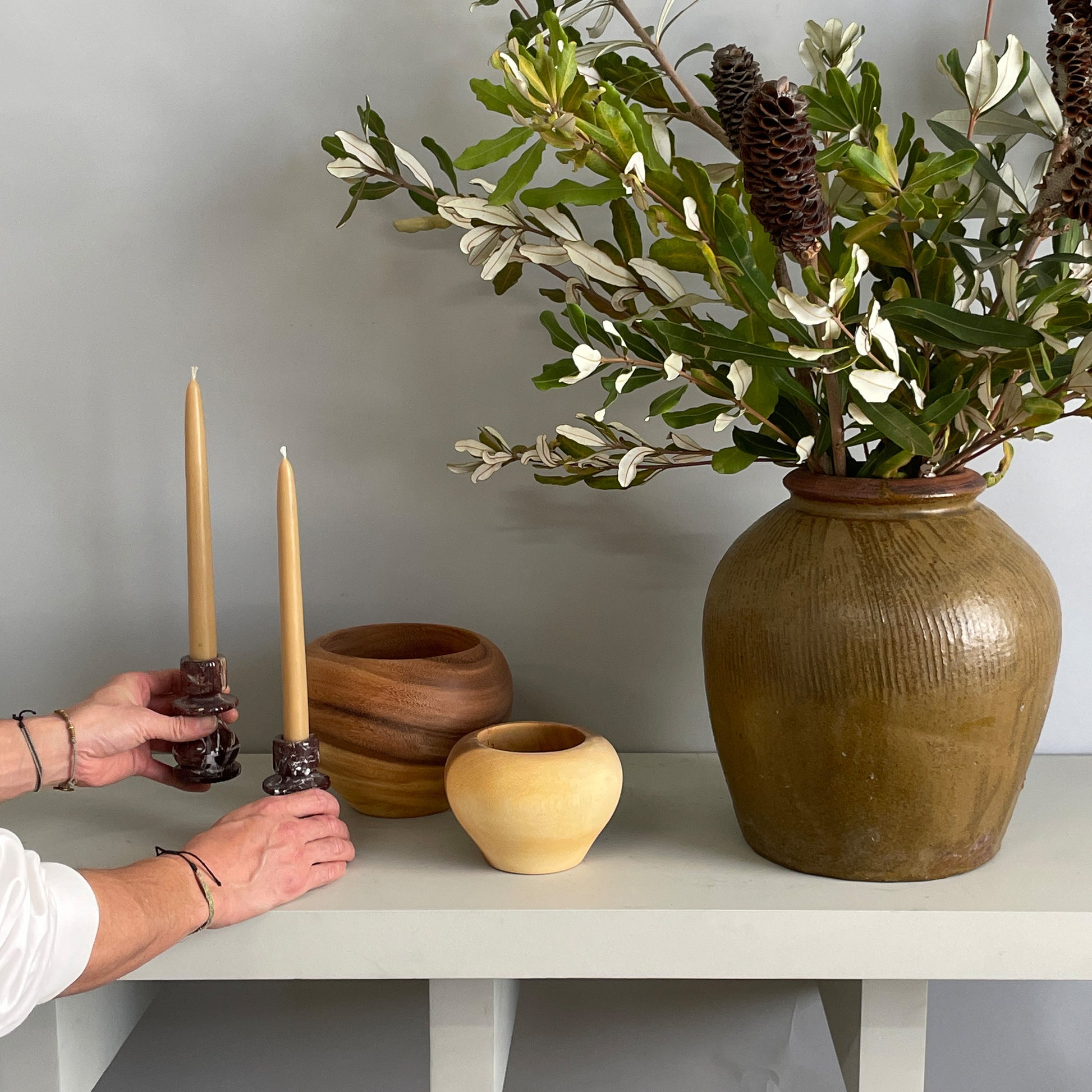 A large olive-green ginger jar with fresh integrifolia foliage styled on a decorative shelf with wooden accents and taper candles.