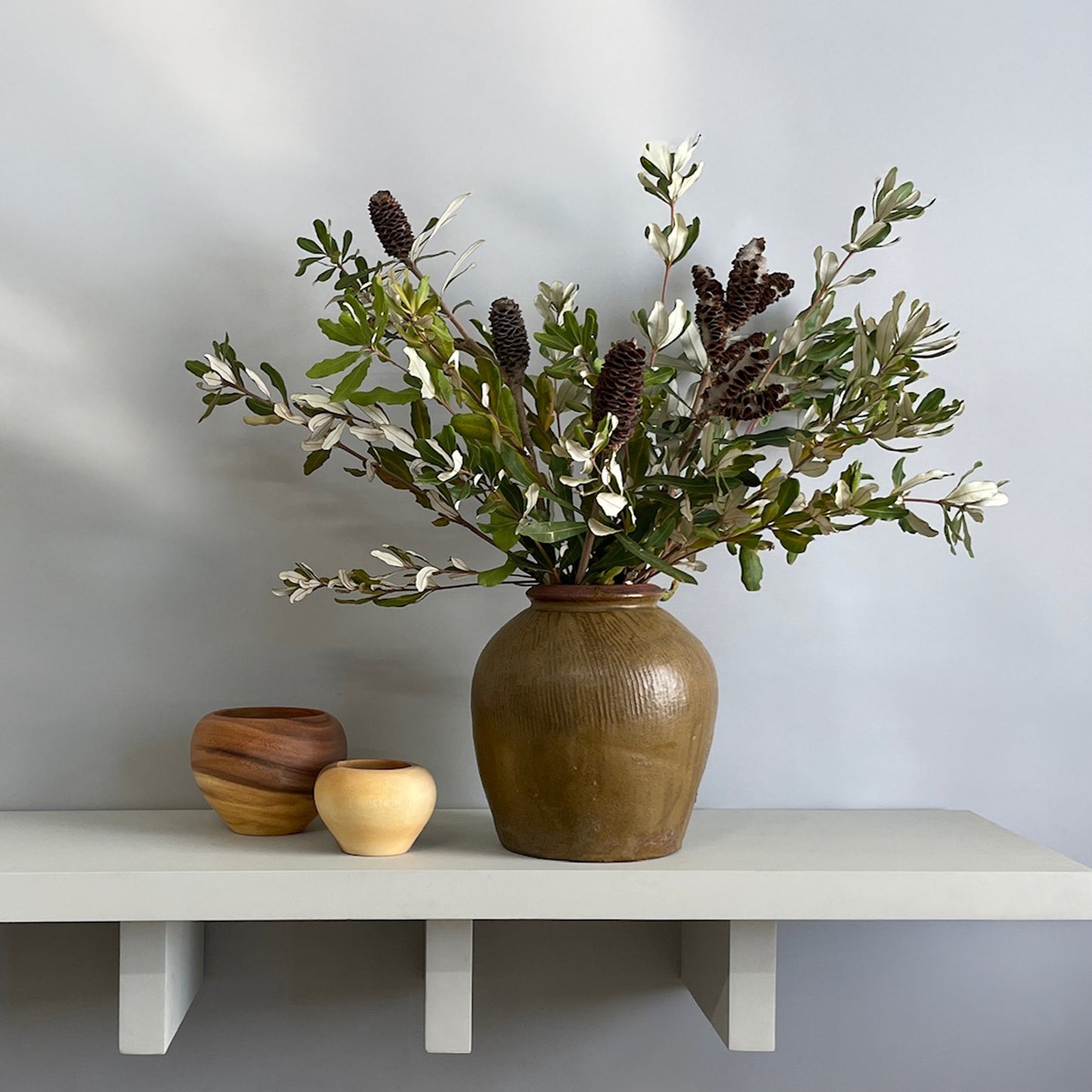 A large olive-green glazed jar with fresh integrifolia foliage styled on a decorative shelf with wooden accents.