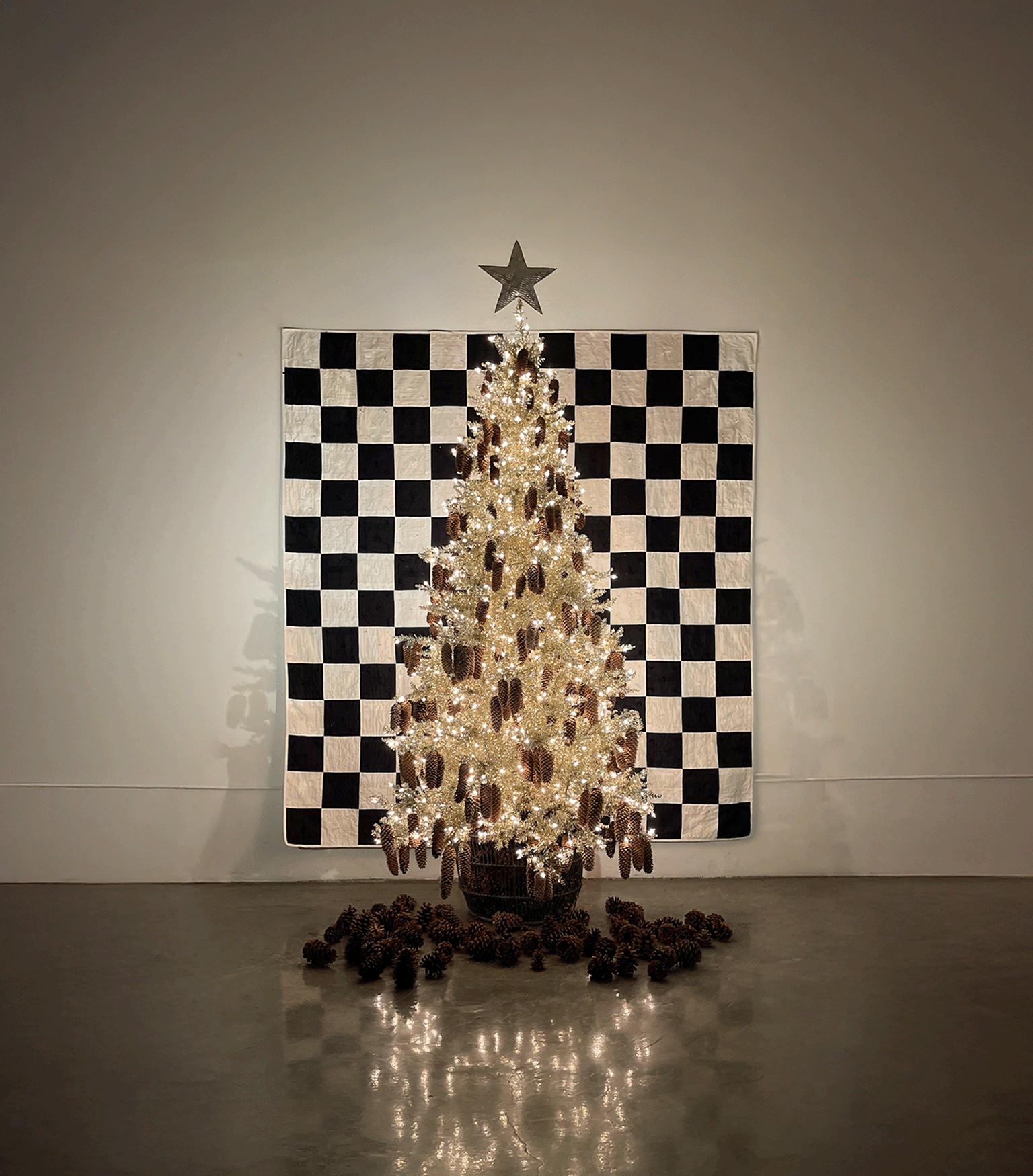 Holiday tree adorned with sparkling lights and pinecones, set against a black-and-white checkered quilt backdrop in a cozy, modern studio space