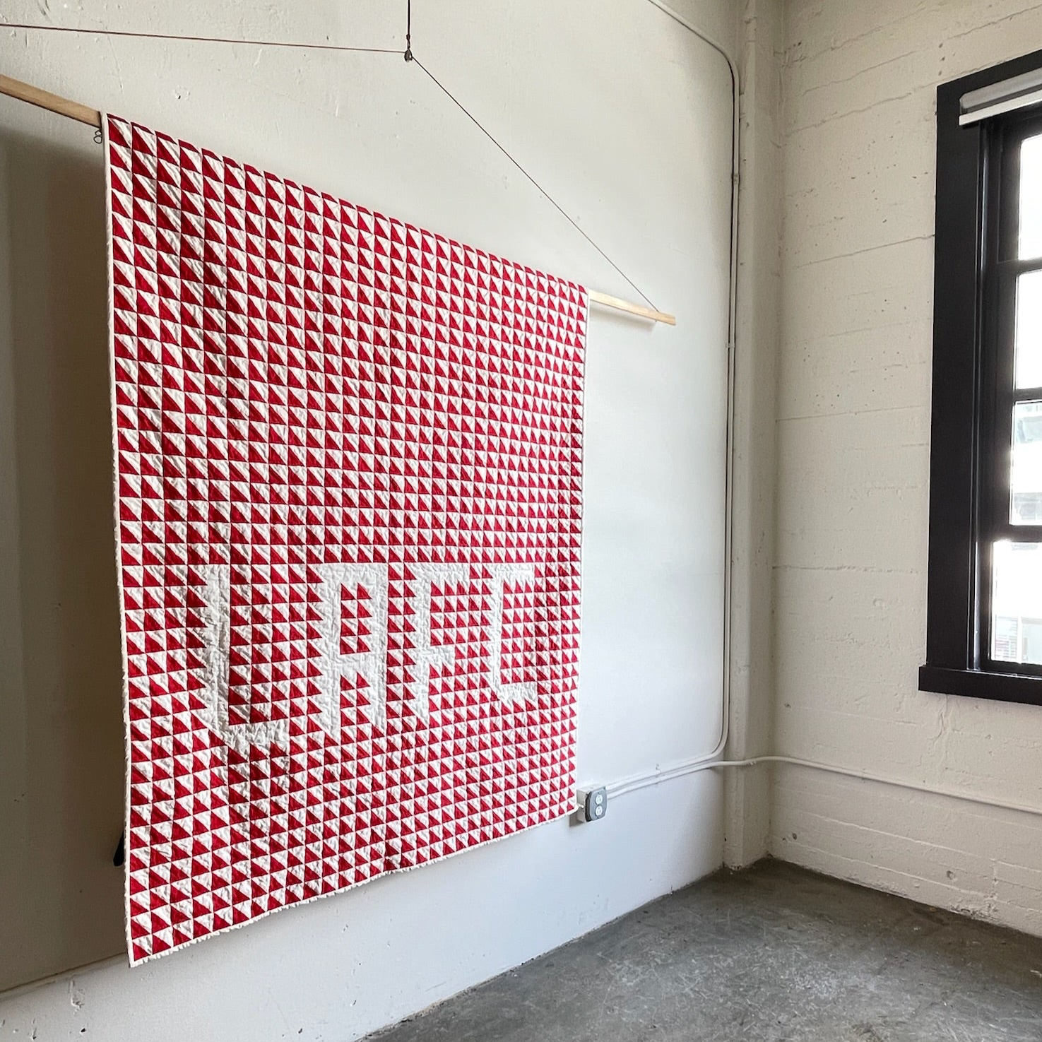 Red and white quilt featuring the letters ‘LAFC’ hanging in a minimalist studio, representing the blend of traditional quilting and contemporary design by Los Angeles Floral Company.