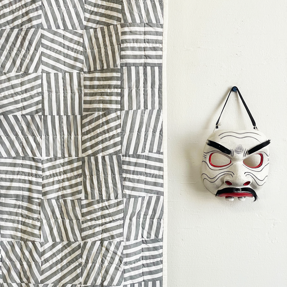 Striped quilt displayed with a traditional Japanese mask, featured on the Wild Card page of Los Angeles Floral Company, showcasing the fusion of American folk patterns and Japanese influences.