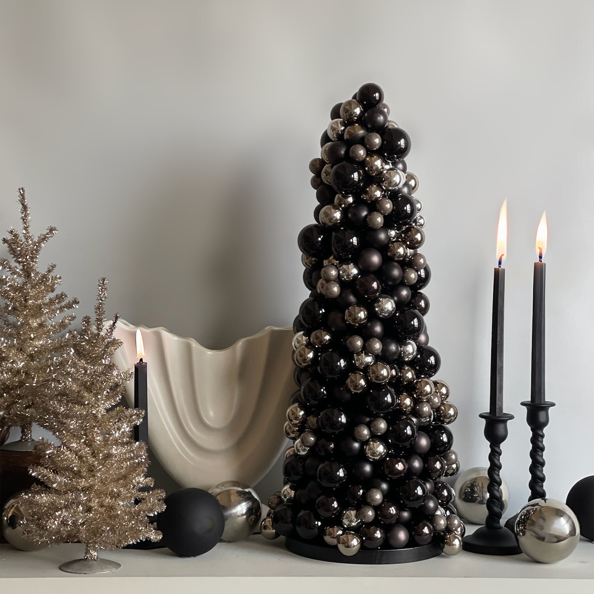 A beautifully styled Moonlit Noir glass bauble cone tree as the centerpiece of a sophisticated holiday display, paired with silver tinsel trees and elegant decor, all resting on a black wood base.