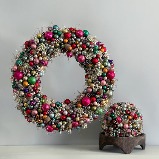 A colorful, oversized wreath made with jewel-toned ornaments, tinsel, snow-dusted pinecones, and natural accents.