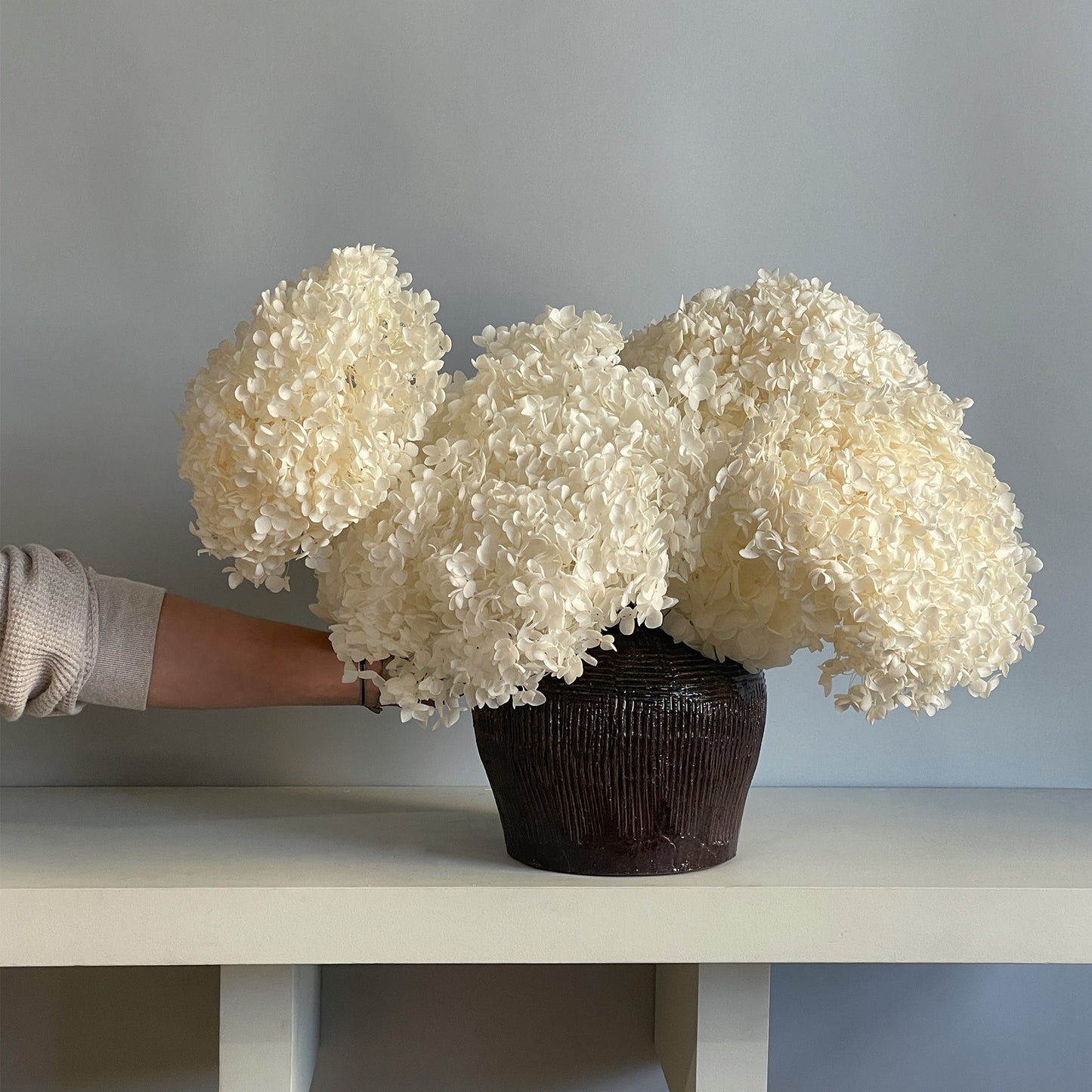 Side view of the preserved white hydrangea arrangement in a brown ceramic glazed jar, highlighting the luxurious, voluminous texture of the flowers.